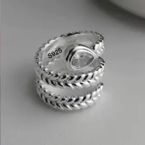 Offener BFCLUB Ring mit geflochtener Ährenstruktur und transparentem Tropfenstein auf grauem Hintergrund