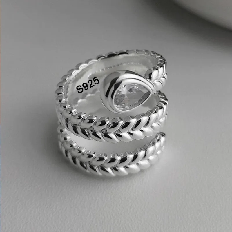 kf-S17131ffeadc14981a17f307701148ab1A Offener BFCLUB Ring mit geflochtener Ährenstruktur und transparentem Tropfenstein auf grauem Hintergrund