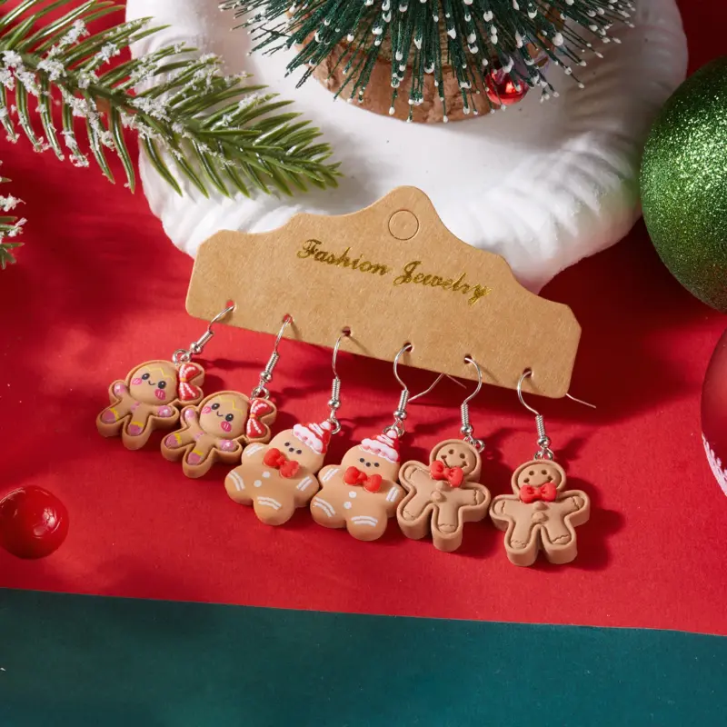 Drei Paar Lebkuchenmann-Ohrhänger mit rosa Schleife, Weihnachtsmütze und roten Fliegen vor roten Dekorationen