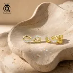 ORSA JEWELS goldfarbene Moissanit Unendlichkeits-Ohrstecker als Paar auf hellem Stein